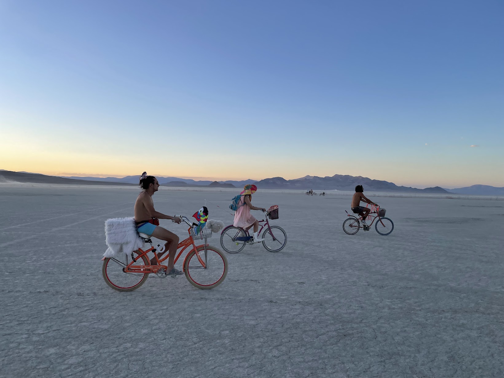 Three riders crossing the open playa at dusk on decorated cruiser bikes — one with a sheepskin on the rack, one in a wide striped sun hat — with distant mountains and an empty pale horizon beyond.