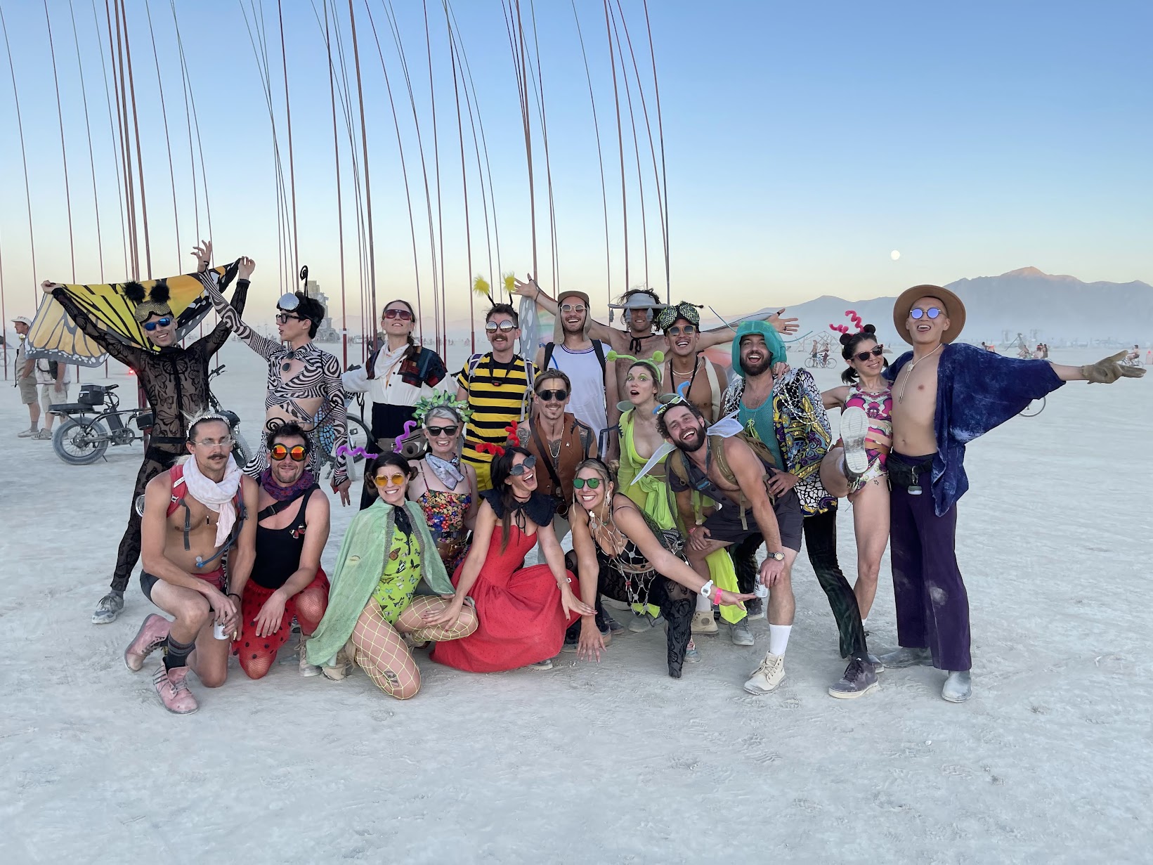 Around twenty camp members posed together on the open playa at dusk, in costume — butterfly wings, bee stripes, antennae, sequins and sunglasses — with bicycles, distant mountains and a rising moon behind them.