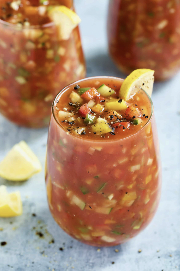 Three glasses of chilled gazpacho, thick with diced tomato, cucumber, pepper and onion, each topped with cracked black pepper and a lemon wedge.